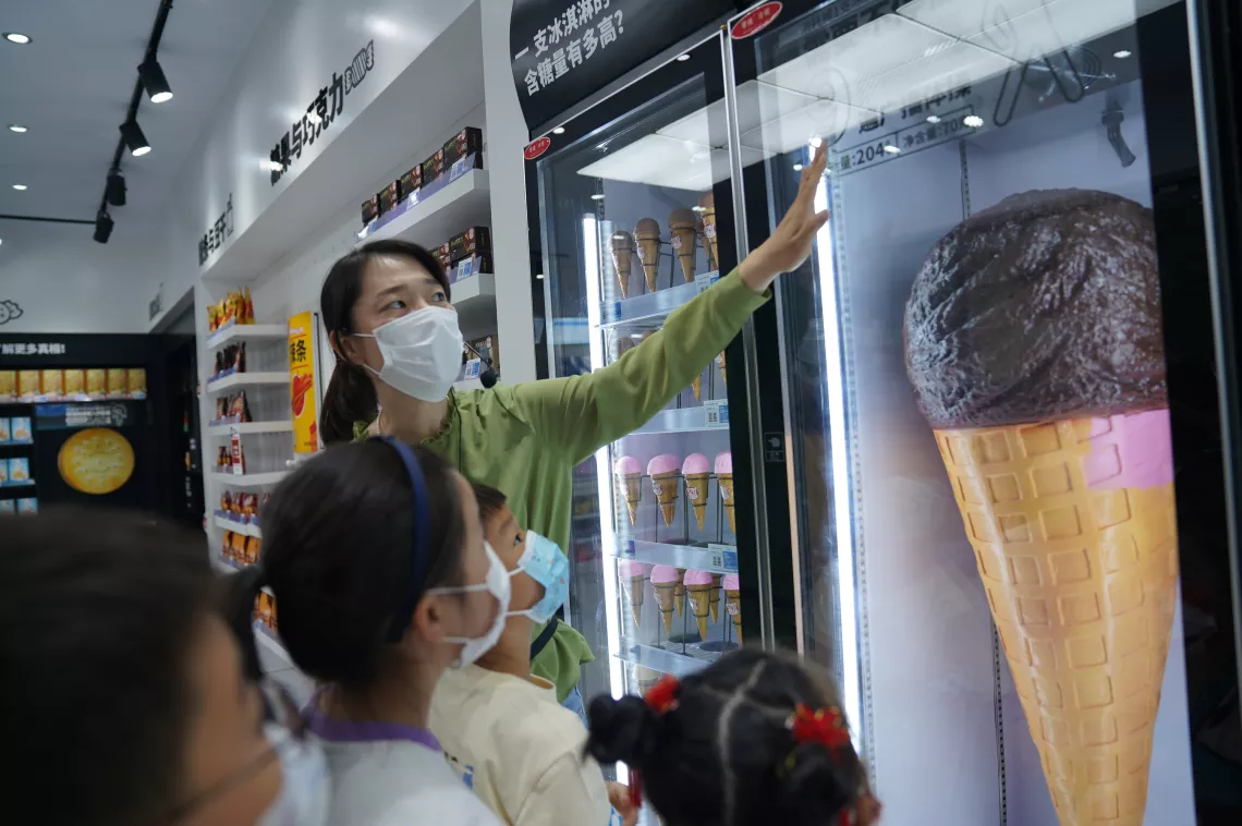 2022年5月21日，在山东省威海市科学技术馆，一名工作人员向参观联合国儿童基金会知食小卖部展览的儿童介绍一支冰淇淋含有多少糖分和脂肪。