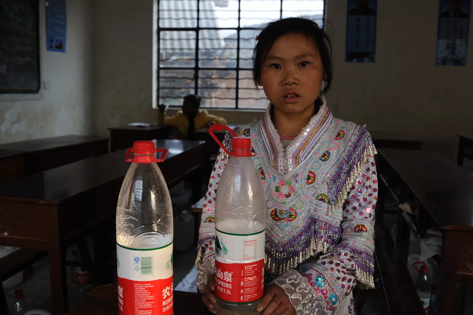 12岁的高小秀在教室内，她旁边放着两个水瓶，装着分配到的水。
