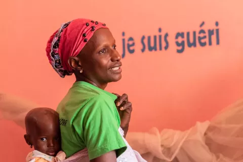 Aïcha stands against a backdrop that says, "I am cured” in French, with Nima on her back.