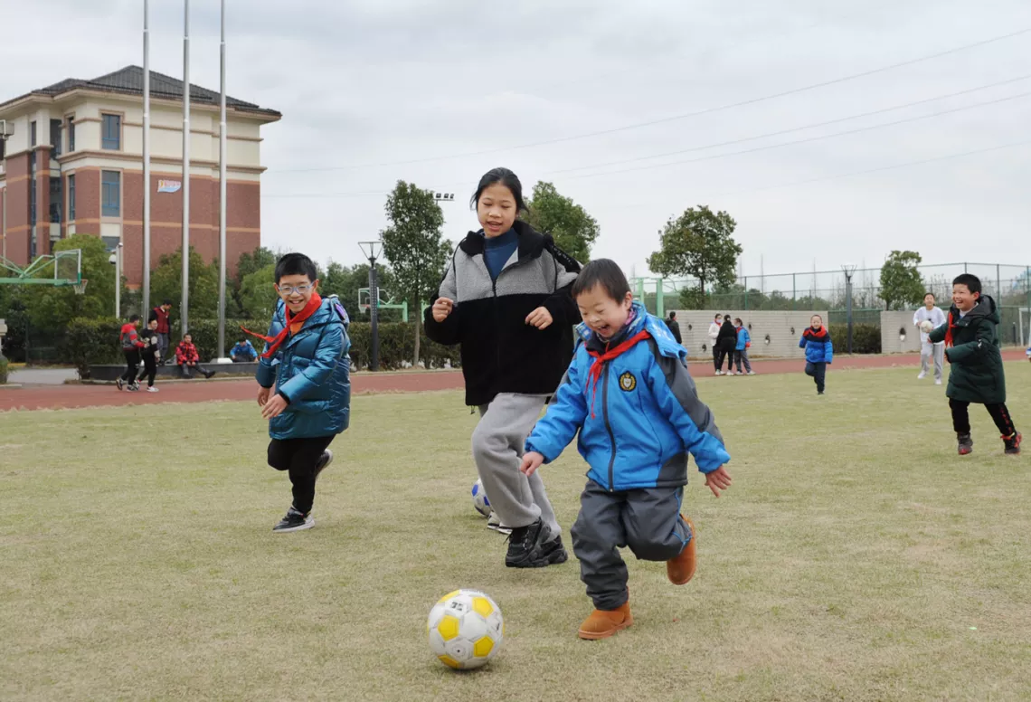校园里的操场上，孩子们在做运动，画面正中是一名戴眼镜的小男孩、一个个子稍高的女孩，以及另一名男孩，在共同追着足球奔跑。