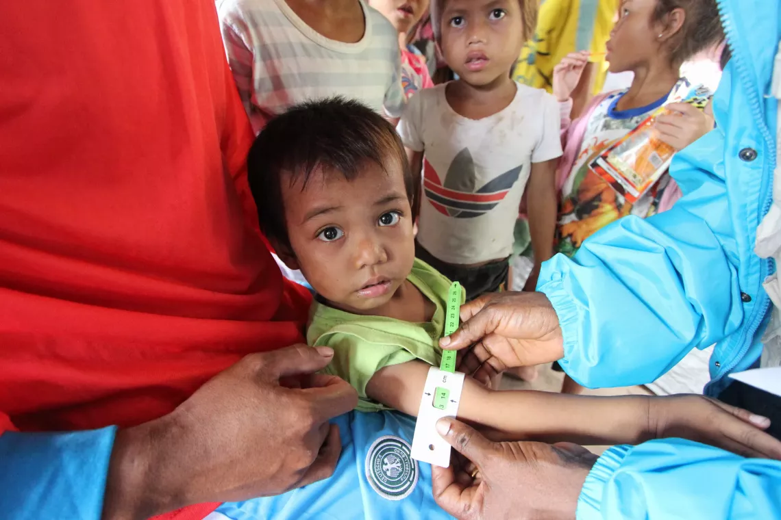 A child stares into the camera as a healthcare worker performs the Mid-Upper Arm Circumference (MUAC) procedure.